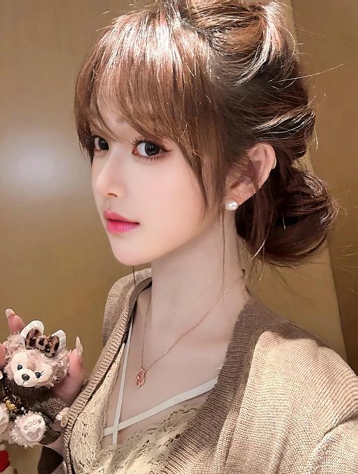 A selfie-style portrait of a young East Asian woman with light brown hair styled in an updo, wearing pearl earrings, a gold necklace with a red pendant, and a beige knit cardigan, holding a small stuffed animal.