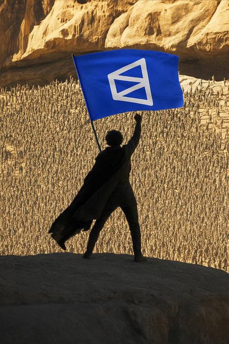 A silhouetted figure in a flowing cloak stands triumphantly on a rocky outcrop, holding aloft a bold blue flag bearing the Polygon ($POLY) logo against a backdrop of sunlit cliffs and an expansive crowd. The image evokes the rallying call to the community, capturing the spirit of unity and action as they kick off a new week of farming, trading, liquidity pooling, and building on the Polygon network.