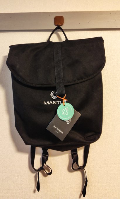 A black backpack with the word "MANTLE" printed on it, hanging on a hook with a teal tag attached.