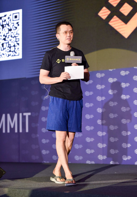 A man wearing a black Binance-branded t-shirt and blue shorts stands on stage at a Binance event, holding a paper and microphone, with a QR code visible behind him.