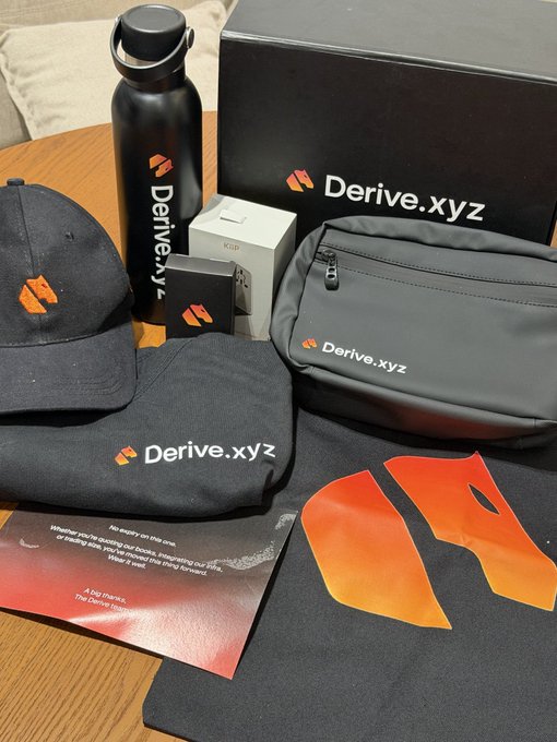 A collection of Derive.xyz branded merchandise including a black baseball cap, water bottle, fanny pack, mouse pad, and packaging box with a thank you card.