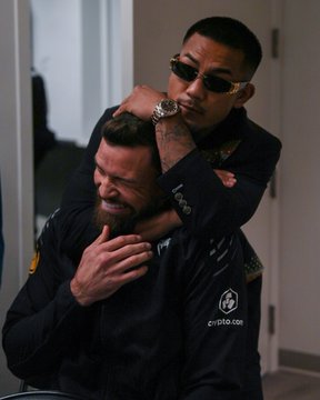 Two fighters, Joshua Van and Ruffy, pose in a mock submission hold indoors, with one applying a rear naked choke to the other as part of a promotional shot. The image features the "crypto.com" logo on the seated fighter's black jacket, tying into the post's #CryptoCom sponsorship reference for UFC 331.