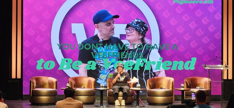 A presentation panel on stage with a person speaking into a microphone, with a large screen behind displaying a photo of two people and the text 'YOU DONT HAVE TO HAVE A VEETFRIENDS to Be a VeeFriend' along with 'PEGACORN 3.0th'.