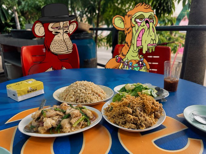 Two cartoon Bored Ape Yacht Club NFTs—one in a black hat and another with melting pink features—are edited as the "diners" at a table laden with plates of stir-fried shark meat, fried mantis shrimp, and a heap of fried rice, alongside a "Bamboe" tissue box and iced drinks.