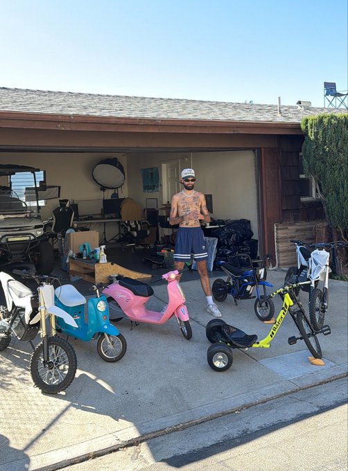 A shirtless man wearing a white cap and blue shorts stands in his open garage surrounded by a collection of small bikes, scooters, and a drift trike, including a pink scooter and a yellow-framed trike with "SILK" printed on it. The scene of assembling various fun, colorful rideables serves as a visual metaphor for his "SOLANA SUMMER" project, suggesting he's building out a vibrant lineup of vehicles.