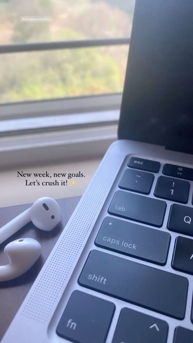 A MacBook laptop sits on a desk next to white wireless earbuds by a window, with overlaid text reading "New week, new goals. Let's crush it! 💪" The motivational workspace image pairs with the post's call to action about getting involved with @FIH_USD1's Web3 prediction markets project this week.
