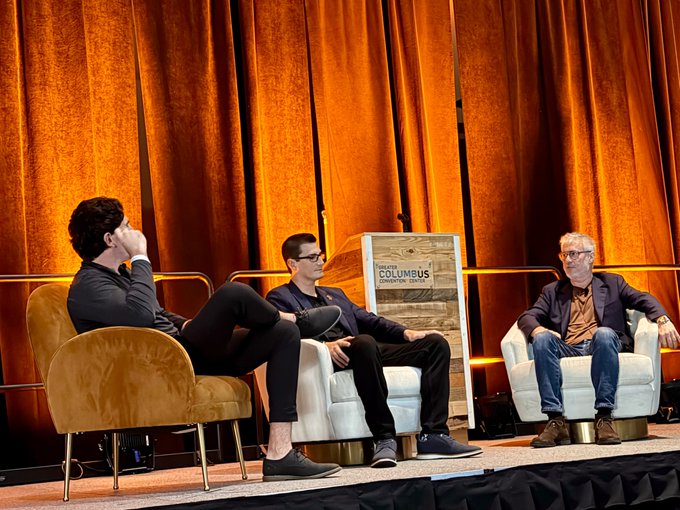 Three men sit on chairs on a stage with orange curtains in the background, with a sign between them reading 'COLUMBUS'.