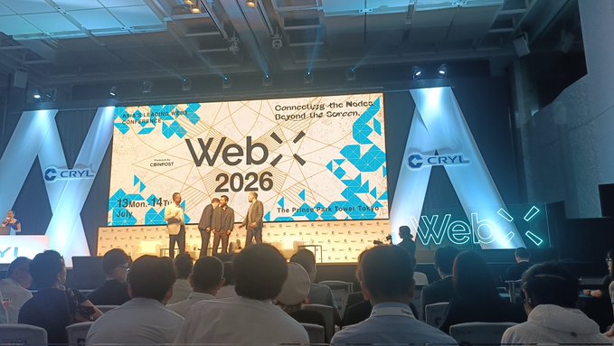 A conference stage with a large screen displaying 'WebX 2026' and 'Connecting the Nodes Beyond the Screen,' sponsored by CRYL, where four presenters are standing in front of a seated audience.