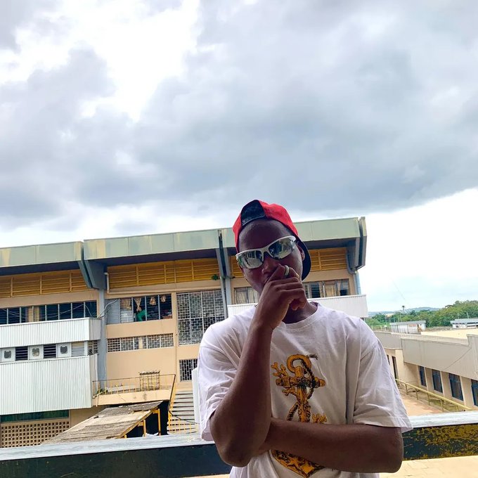 A young man wearing a red cap backwards, sunglasses, and a white t-shirt standing on a rooftop with a school building behind him.