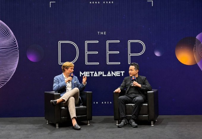 Two men seated on black chairs on a stage having a discussion at 'THE DEEP' event hosted by METAPLANET in Hong Kong, with a dark blue cosmic-themed backdrop.