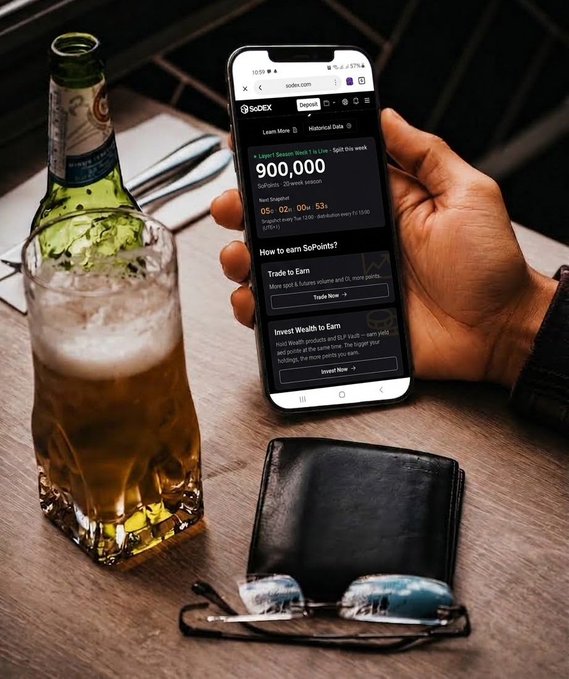 A hand holding a smartphone displaying the SoDEX platform with 900,000 SoPoints for the Layer3 Season Week 2, sitting next to a glass of beer and a wallet with sunglasses on a table.