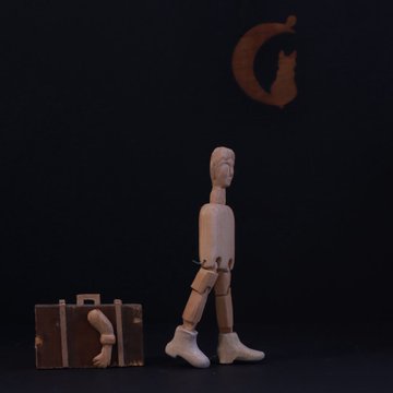 A wooden articulated mannequin figure in white boots walks past a small suitcase containing a smaller figure, beneath an orange crescent moon shape against a black background.
