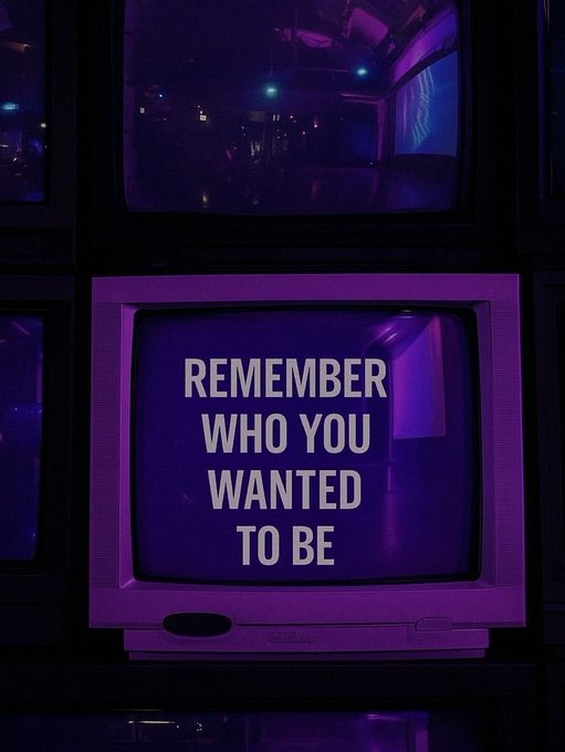 An old CRT television displays the text "REMEMBER WHO YOU WANTED TO BE" in white letters on a purple background, surrounded by other screens in a dark, neon-lit space.