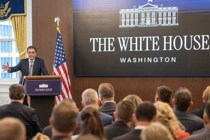 A man in a suit speaks at a podium labeled "THE WHITE HOUSE" with a large screen behind him displaying "THE WHITE HOUSE WASHINGTON," addressing a seated audience in the briefing room. The image visually supports the post's theme of the U.S. executive branch driving its "American Drone Dominance" agenda, as the speaker delivers an official policy announcement from the White House press room.