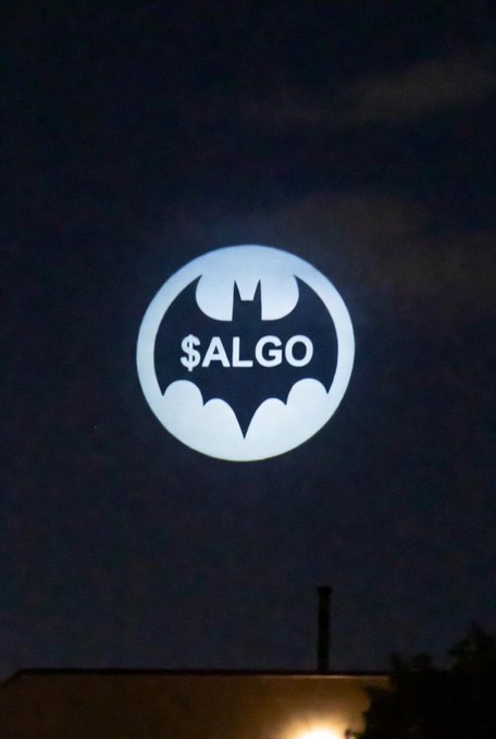 In the image, there is a large white circle with a black bat inside of it that has the word "ALGO" written on it. The bat is illuminated against a dark sky at night, creating an interesting visual effect. The caption below the image reads "The Bat Signal is FLASHING ALGORAND $ALGO". This suggests that there might be some sort of event or promotion related to the bat and its symbolism in the context of the post.