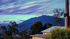 A scenic view from a hilltop or elevated terrace shows distant blue mountains and a tall communications tower under a cloudy blue sky, with rooftops visible in the foreground.
