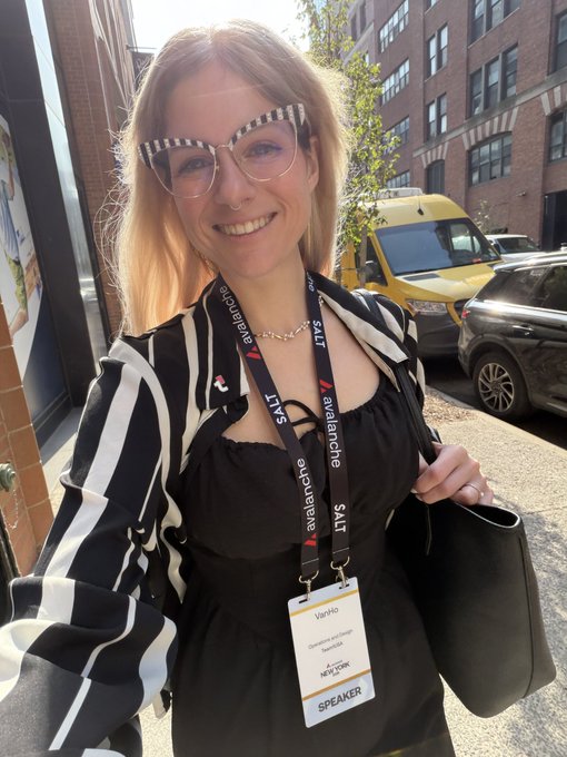 A smiling woman with cat-eye glasses and blonde hair wears a black and white striped top with an 'Avalanche' lanyard reading 'SALT' and a 'SPEAKER' badge, holding a black bag on a city street.