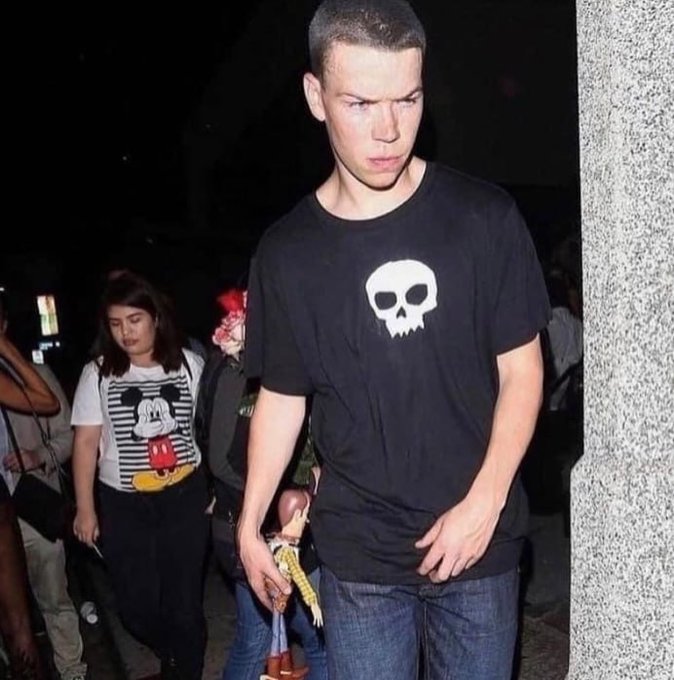 A photo of a young man with a buzz cut wearing a black t-shirt with a white skull design, standing in a crowded nighttime setting with other people visible in the background.