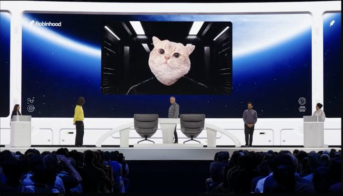 A Robinhood event stage features a massive screen displaying a cat's face, with presenters standing at the front while a crowd watches from the audience. The Robinhood logo is visible in the upper left corner, connecting to the post's claim that the platform is shilling a "cat" themed token.