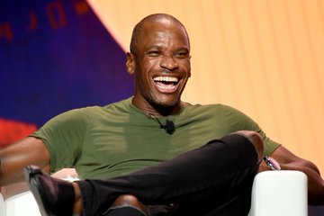 iced coffee on a table, with a man in a green shirt and black pants seated at the table, smiling warmly.