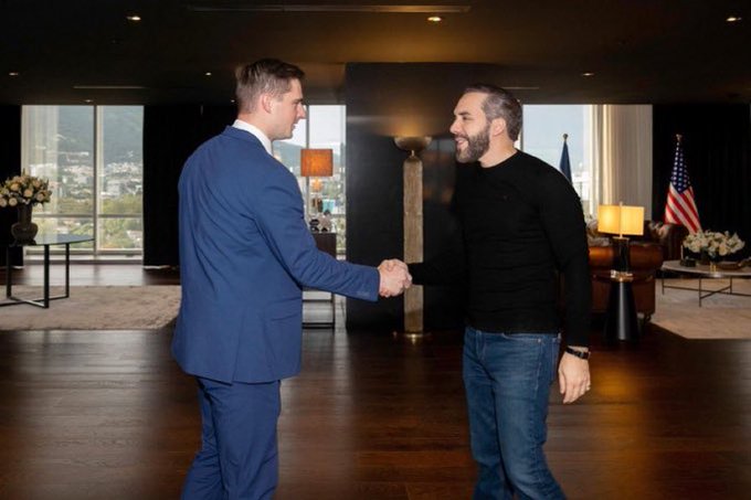 Two men shake hands in a formal interior room with wood floors; one wears a blue suit and the other a black sweater, with American flags visible in the background.
