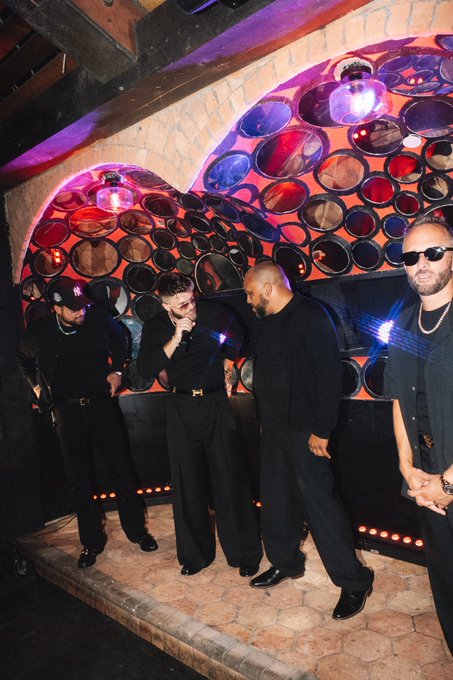 Four men dressed entirely in black—including sunglasses, a black cap, and chains—stand closely together in a dimly lit, upscale venue featuring an ornate arched wall decoratively stacked with red, blue, and black circular elements that resemble wine barrels or discs, illuminated by hanging pendant lights; the accompanying post "Can I get a GM?" suggests a playful morning greeting to their online followers, while the image likely captures a night out, possibly at a stylish bar or club, with the group's coordinated all-black outfits and confident poses reinforcing a bold, nightlife-ready aesthetic that contrasts humorously with the upbeat morning message.