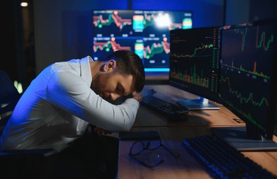 This image shows a man lying down next to his desk with a computer monitor displaying stock market charts. The man appears to be sleeping or resting during work hours. The background includes another computer screen, suggesting that this workspace may belong to him, possibly connected to different tasks or responsibilities at hand. A caption in the image reads: "I watched SOL go from 
$18 to triple digits after selling my entire bag and still haven'n forgave myself @RallyOnChain". This seems like a somewhat stressful situation for the man as he is struggling with his finances while trying to maintain or regain control of his investments. The presence of multiple screens might indicate that this work environment requires multitasking, with several tasks demanding attention simultaneously.