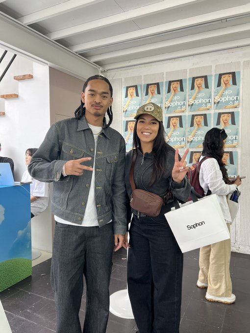 A man and a woman pose together indoors, both flashing peace signs, with the woman wearing a Sophon cap and carrying a shopping bag labeled "Sophon," in front of a step-and-repeat banner featuring "Sophon" branding.