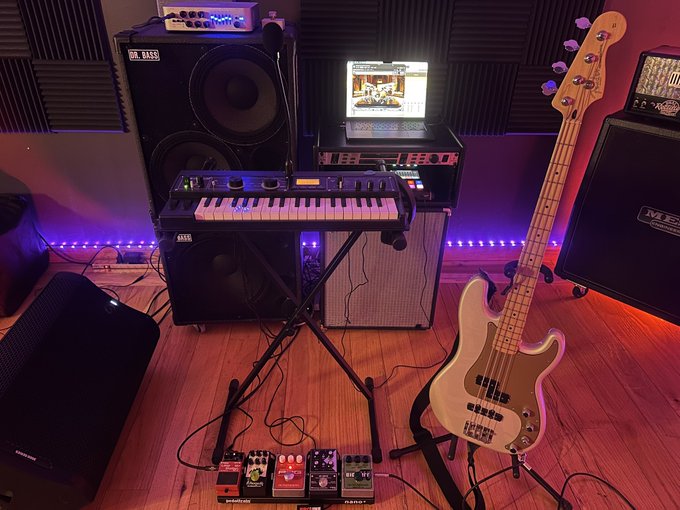 A home music studio setup features a bass guitar on a stand, a keyboard/synth, an amplifier, an Orange bass cab, a laptop, and several guitar effect pedals on the floor, all illuminated by purple LED lighting.