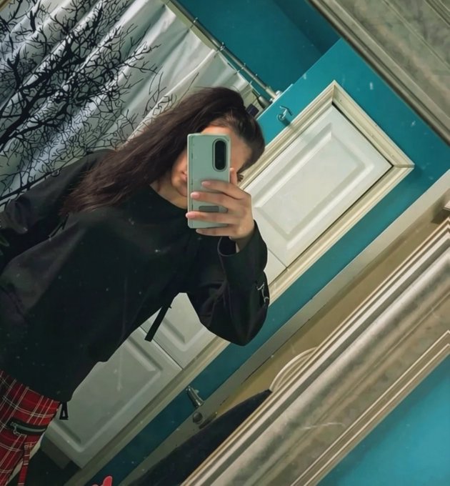 A young woman with long dark hair takes a mirror selfie in a bathroom with teal walls and a marble-framed mirror, holding a light green smartphone while wearing a black jacket and red plaid pants.