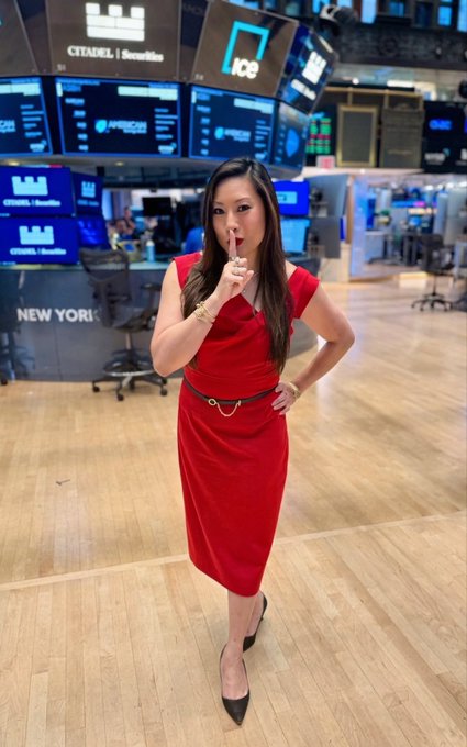 A woman in a red dress with a gold belt stands in what appears to be the New York Stock Exchange trading floor, with screens displaying 'CITADEL Securities', 'ICE', and 'NYSE' in the background.