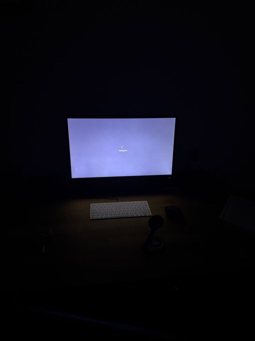 A dark room where the only visible elements are a computer monitor displaying a faint Samsung boot/loading screen, a white keyboard, and a mouse on a desk.