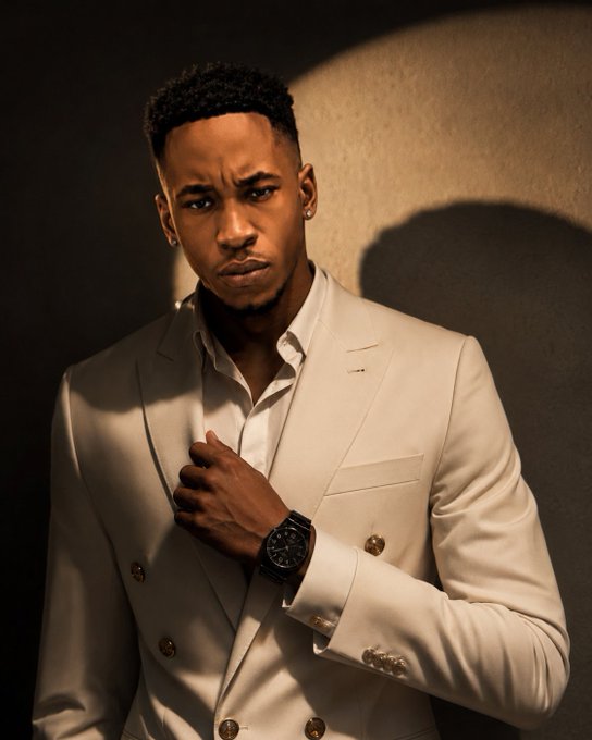 A portrait of a young man in a cream-colored double-breasted suit and white shirt, wearing a luxury wristwatch, posing against a beige wall with dramatic lighting.