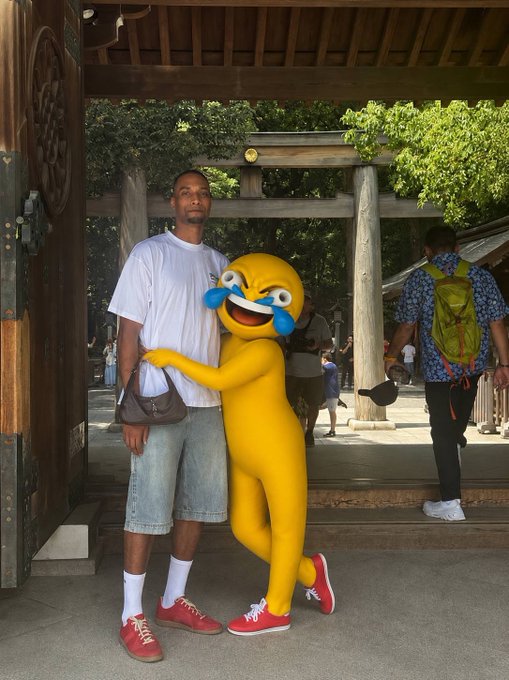A man poses with a person in a yellow laughing emoji costume near a wooden torii gate at a Japanese shrine, while another visitor with a green backpack walks by.