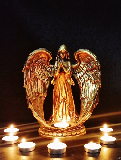 A golden angel figurine with large outspread wings stands illuminated against a black background, surrounded by several lit tealight candles at its base.