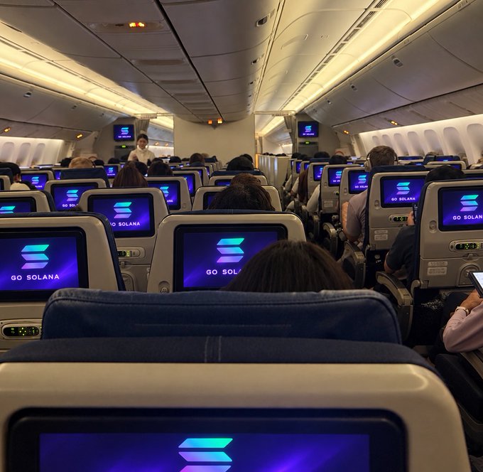 The interior cabin of a commercial airplane with passengers seated, where every seatback screen displays the 'GO SOLANA' logo and branding.