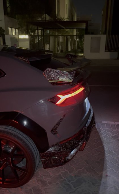 A rear view of a customized dark-colored sports car parked at night, featuring a large spoiler, carbon fiber accents, and the word "Venatus" on the rear.