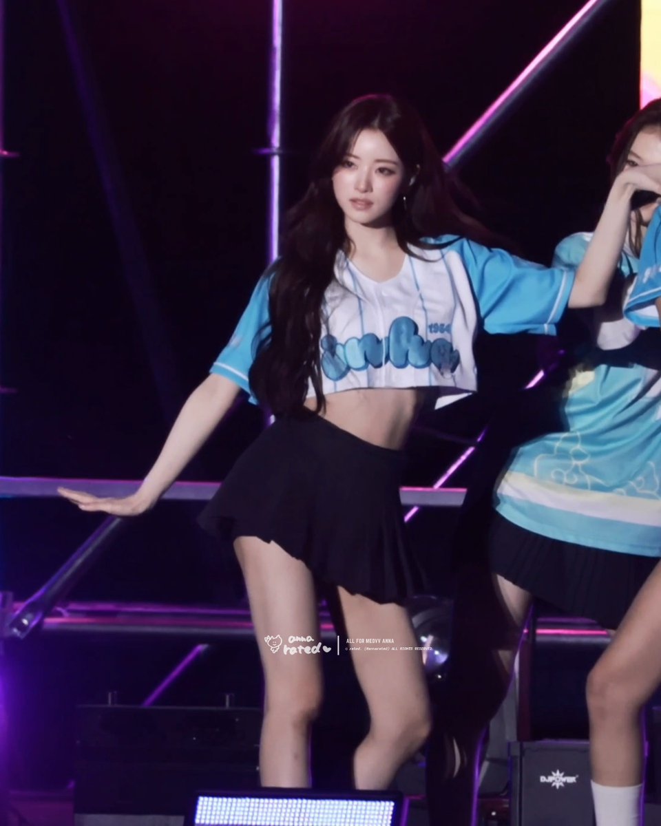 A K-pop idol with long dark hair performs on stage wearing a blue and white baseball-style jersey and black pleated skirt, with watermark text 'ALL FOR MEDIV ANNA' visible.