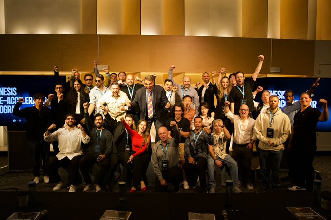 A large group of approximately 25 people pose enthusiastically with raised fists on a stage at what appears to be "Genesis Re-Acceler" or "Re-Accelagr" event.