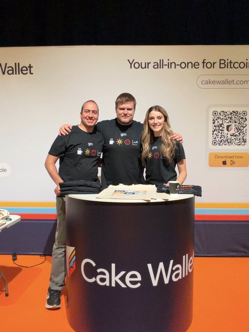 Three people in matching black "gm" t-shirts pose behind a circular "Cake Wallet" booth at what appears to be a trade show, with a backdrop reading "Your all-in-one for Bitcoin" and a QR code.