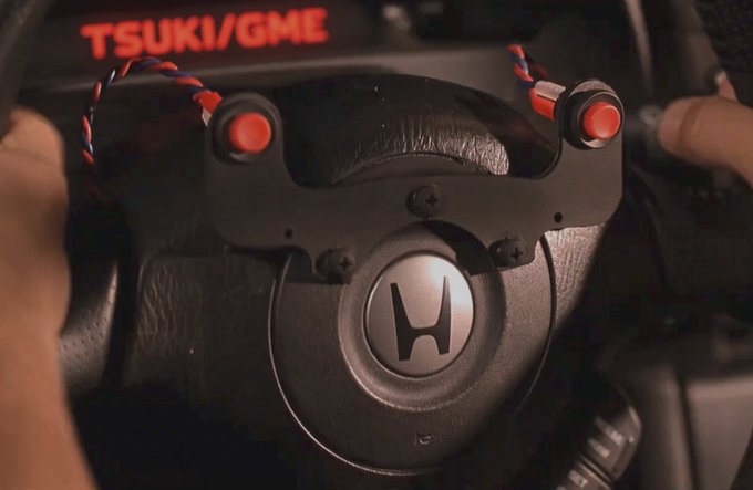 A close-up view of a Honda steering wheel with a TSUKI/GME button module installed, featuring red buttons and colored wiring.