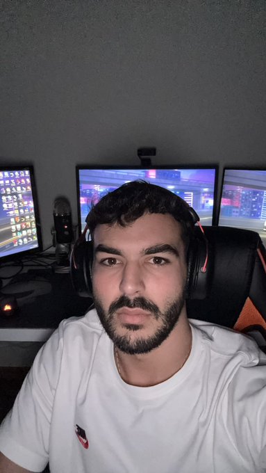 A man with a beard wearing headphones and a white shirt, sitting in a gaming/streaming setup with multiple monitors displaying charts in the background.