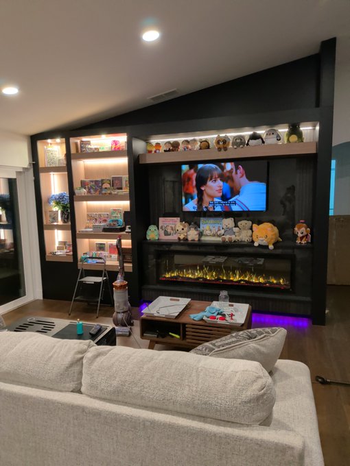 Cozy living room with vaulted ceilings, featuring a built-in entertainment wall with shelves displaying plushies, a TV showing a show, and an electric fireplace beneath, with a beige couch in the foreground.