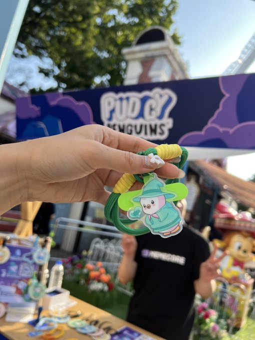 A hand holds a green and yellow rubber charm featuring a cartoon penguin character, with 'PUDCY PENGUINS' signage visible in the background.