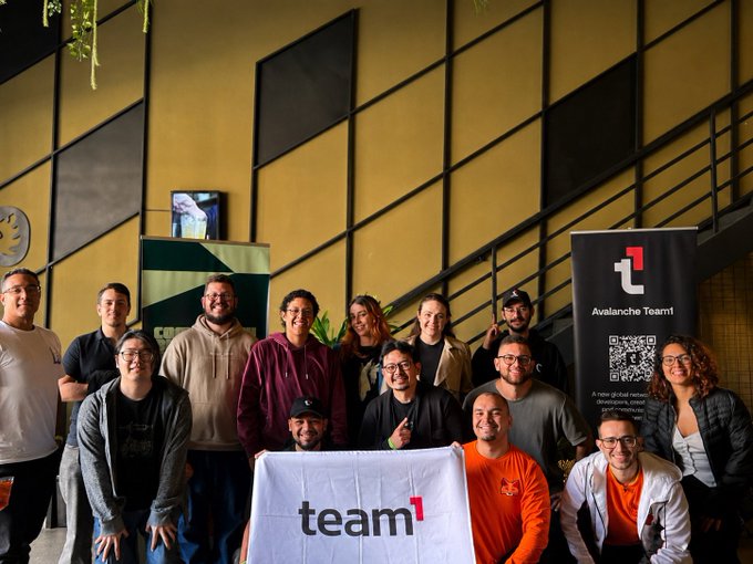 The image features a group of people posing together for a photo with a large white banner that says "Team" in black letters. The team members are standing close to each other, creating an atmosphere of camaraderie and unity. In addition to the banner, there is also a smaller banner visible on the right side of the image.

The background includes a staircase, which adds depth to the scene and suggests that this gathering might be taking place in a multi-level building or event space. The people are standing near potted plants placed at various positions within the room, adding some greenery to the setting.