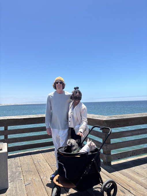 A couple posing on a sunny pier by the ocean with two French bulldogs in a black pet stroller, the man wearing a yellow beanie and the woman wearing a beige jacket and sunglasses.