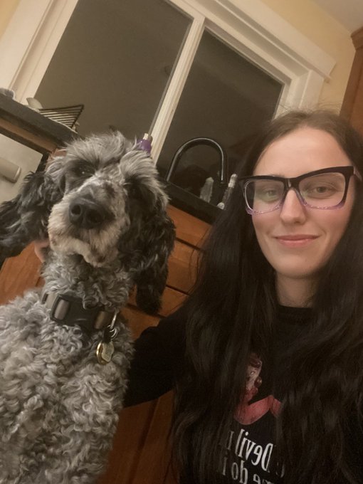 A selfie of a woman with long dark hair wearing purple glasses and a black graphic t-shirt, sitting next to a curly gray dog wearing a harness.