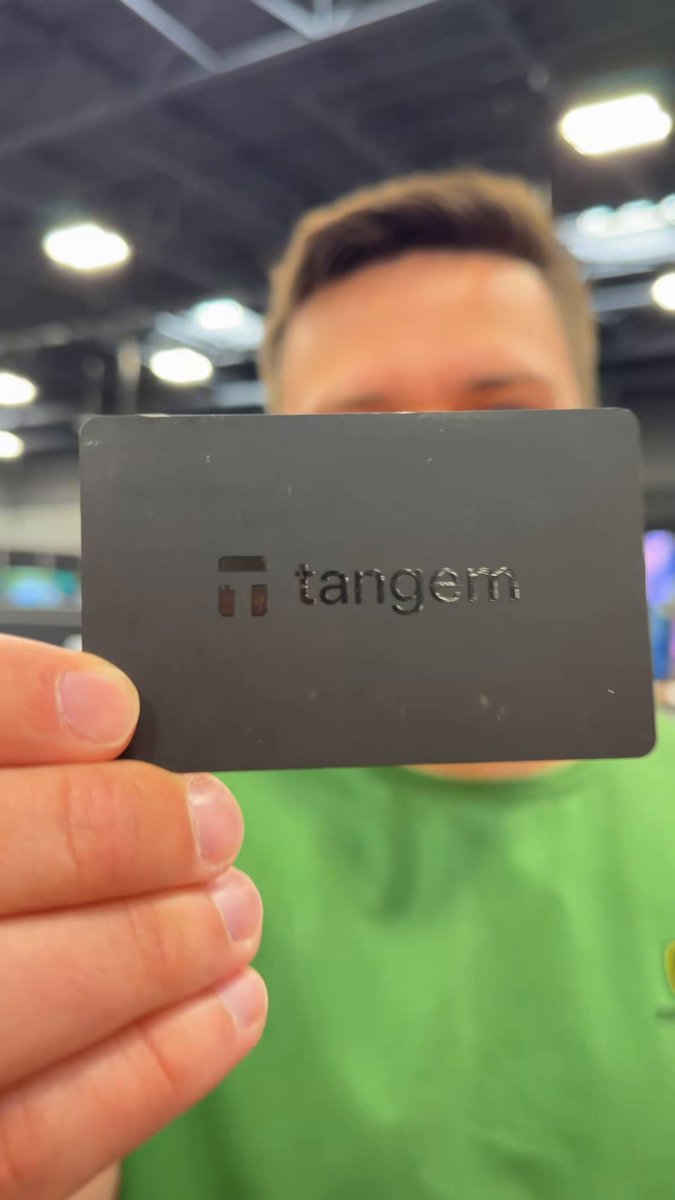 The image features a man holding up a black credit card with the word "Tangem" written on it. The credit card appears to be from Best Buy, as indicated by the text in the image. The man is wearing a green shirt, which contrasts nicely with the black background of the credit card and the post. The caption reads: "Just stopped by Best Buy and saw something I never thought I'd see a few years ago... A @Tangem hardware wallet sitting on the shelf of a major US retailer. I've been using Tangem for over 2 years, so seeing them reach this point is pretty incredible."