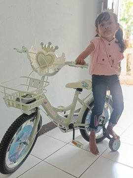 A young girl in a pink sweater vest and jeans stands barefoot with a decorative white bicycle indoors.