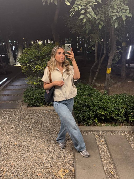A woman with long blonde hair takes a mirror selfie at night while wearing a beige zip-up shirt, blue jeans, and silver sneakers, carrying a black shoulder bag.
